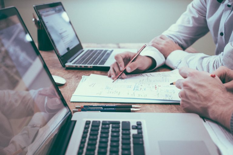 Side view of two people siting next to each other at a desk. There is a laptop in front of each person, and a stack of papers between this. The only part of the people that is visible is their hands, one of which is writing something on the stack of papers.